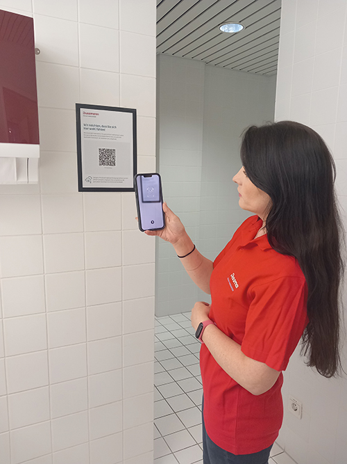 Eine Reinigungskraft in roter Arbeitskleidung Steht in einem Sanitärraum und scannt mit einem Smartphone den QR-Code auf einem an der Wand hängenden Sanitärraumzettel, indem sie die Smartphone-Kamera auf den Zettel richtet.