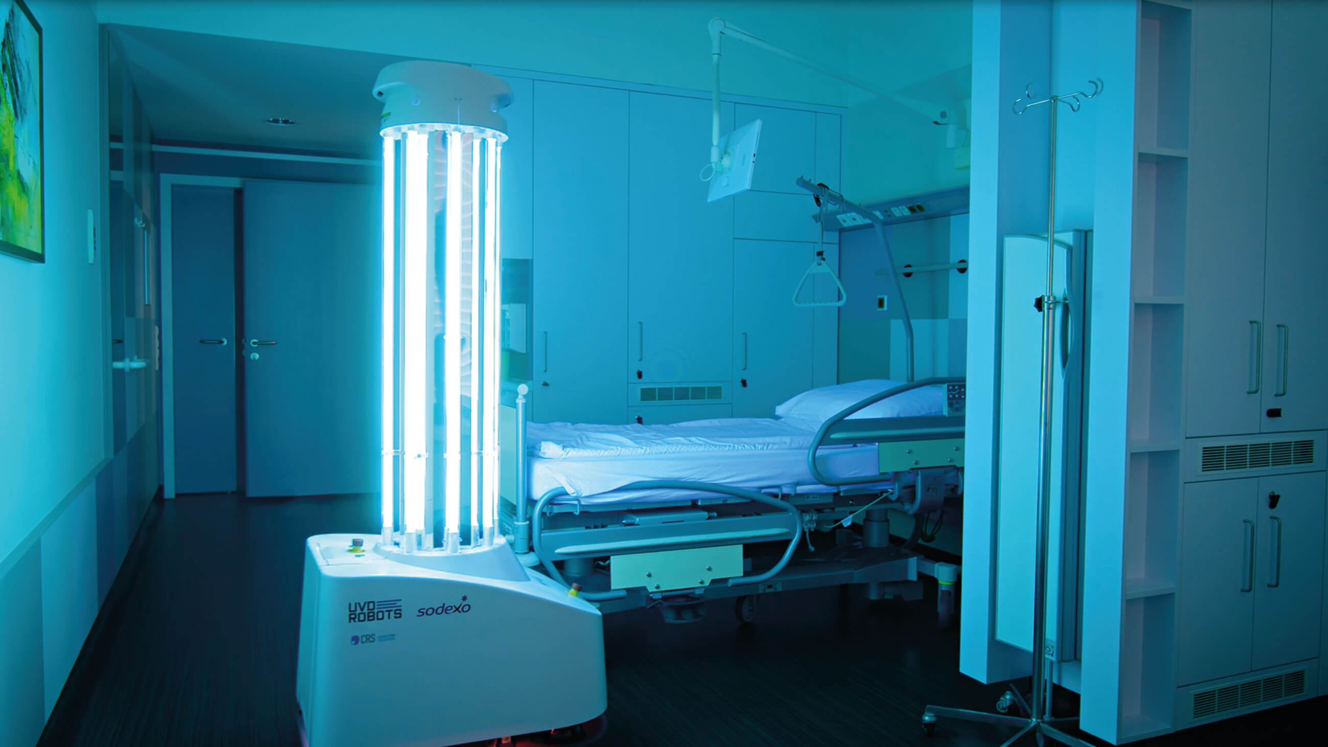 Ein UV-C-Roboter in einem Krankenhauszimmer. Der ungefähr menschengroße Roboter besteht aus säulenförmig angeordneten, leuchtenden Röhren, die auf einem etwas breiteren mobilen Sockel mit der Aufschrift "UVD Robots – Sodexo" stehen. Der Raum ist in türkis-blaues Licht getaucht.