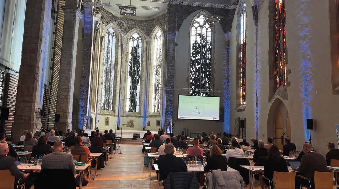 Die Johanniskirche in Magdeburg während des IKS-Forums 2022. Anstelle von Kirchenbänken stehen im Mittelschiff Konferenztische und -stühle, an denen Personen sitzen. Vor den Altarraum ist rechts vorne eine Leinwand angebracht. Vor der Leinwand steht ein Redner an einem Rednerpult.